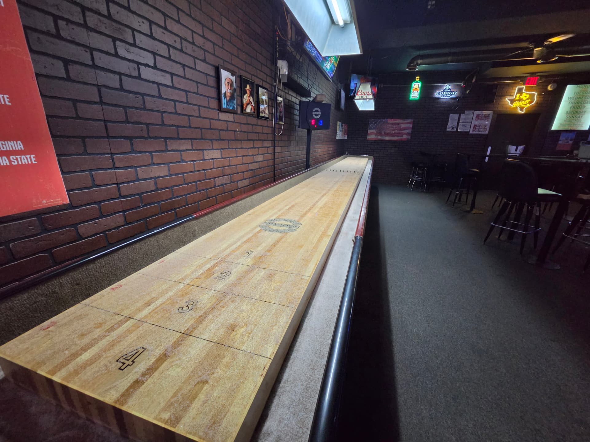 The shuffleboard table along the brick wall with the electronic scoreboard