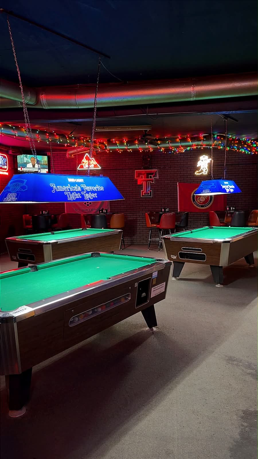 Three green-felt pool tables under Bud Light lamps with Christmas lights and a Texas Tech sign