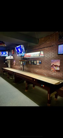 Long shuffleboard table against the brick wall under a Coors Light lamp